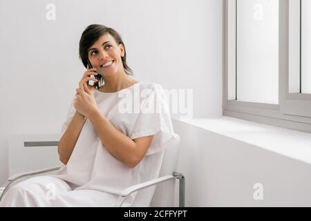 Cheerful Woman in white robe sitting on bed near bed and using smartphone in ward of modern clinic Stock Photo