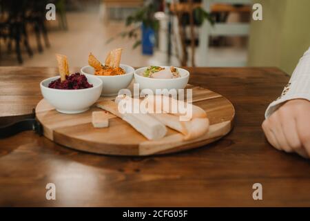 From above crop unrecognizable hands grabbing assorted vegetable dips with crispy crunchy bread in bowl on round stand on table Stock Photo