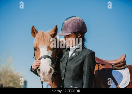 Side view of young teen Woman in jockey helmet and jacket caressing horse standing together outdoors against blue sky Stock Photo