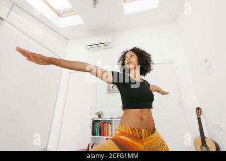 African American attractive young Woman performing yoga posture with stretched arms on mat in light room