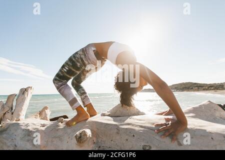 Side view of African American attractive young Woman standing in yoga posture like bridge in open air on background of calm water in sunny day Stock Photo