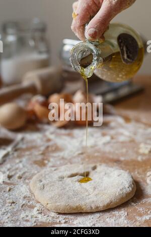 faceless cook pouring fragrant fresh olive oil from glass jug onto rolled pasta dough on background of blurred kitchen mess Stock Photo