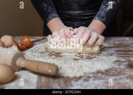 unrecognizable housewife in black clothes kneading elastic dough with flour on table with hands while preparing pastry in cozy kitchen at home Stock Photo