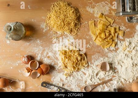 Top view of raw different types of pasta locating on messy wooden table covered with flour with bottle of olive oil eggshells and kitchen utensils Stock Photo