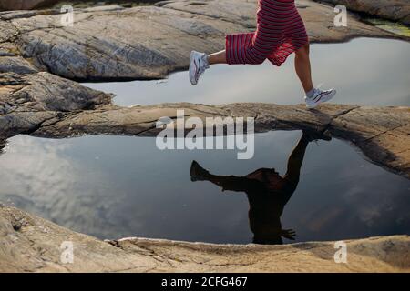 Side view of anonymous crop female traveler in white sneakers and skirt running between puddles on rocky ground on sunny day Stock Photo