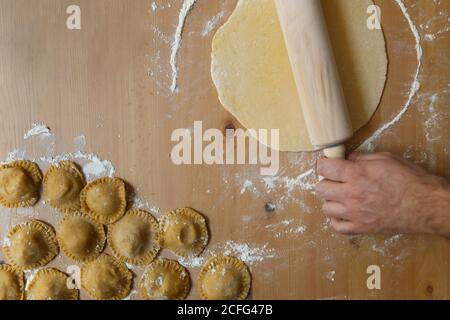 Top view of anonymous person rolling soft dough by rolling pin on table with flour near raw round delicious ravioli in kitchen Stock Photo