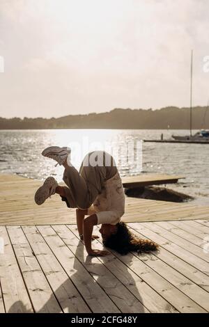 Side view of female dancer in casual clothes and white sneakers standing on head on wooden boardwalk on sunny day during vacation Stock Photo