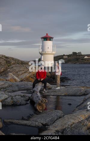Adult man in red jacket sitting on fallen tree trunk and Woman in casual clothes standing on stones near river and lookout tower looking at camera on cloudy day Stock Photo