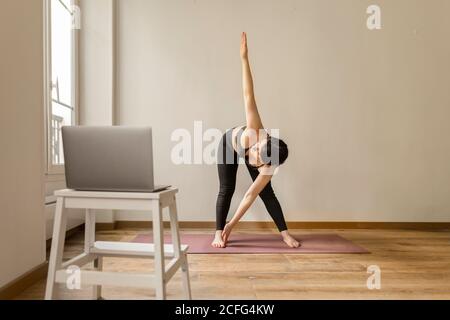 Side view of female in sport clothes standing in Adho Mukha Svanasana while practicing yoga on mat at home near stool with laptop Stock Photo