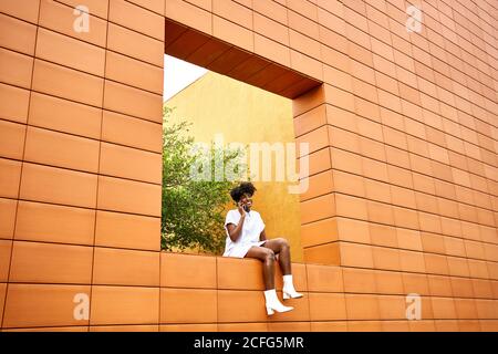 Low angle of young African American female in trendy white outfit smiling away and making phone call on street while sitting on wall contemporary building with bright orange and yellow walls Stock Photo