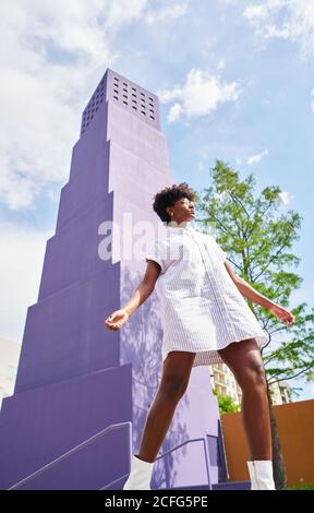 From below view of slender African American Woman in white dress and boots posing alone against multistory violet building in city in sunny summer day Stock Photo