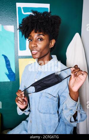 Positive young African American female in casual wear looking at camera demonstrating black protective mask for coronavirus prevention while standing near wall in modern room at home Stock Photo