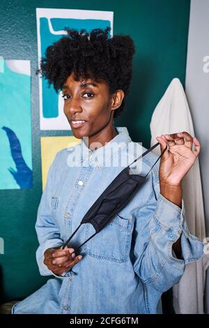 Positive young African American female in casual wear looking at camera demonstrating black protective mask for coronavirus prevention while standing near wall in modern room at home Stock Photo