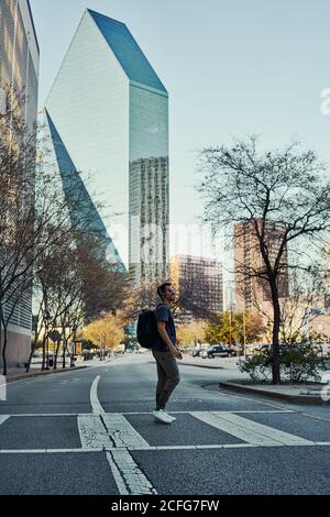 Happy hispanic male traveler in casual outfit and sunglasses with backpack with on headphones while sightseeing at downtown Stock Photo
