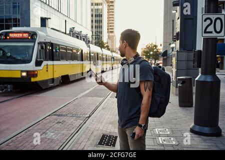 Anonymous Hispanic man in casual wear with backpack surfing on mobile phone while standing on platform at city street Stock Photo