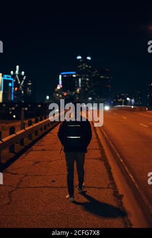 Back view of anonymous man in casual black leather jacket with backpack walking at night city on blurred background