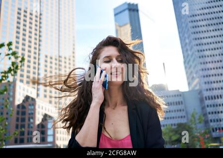 Content businessWoman in elegant jacket discussing work issues while speaking on cellphone and standing on blurred background of skyscrapers Stock Photo