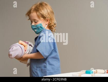 young child wearing medical PPE holds a swaddled baby doll Stock Photo