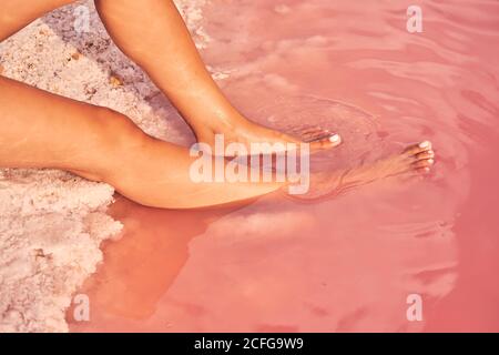 Crop side view of bronzed Woman relaxing on red lagoon shore with legs in pink salty water Stock Photo