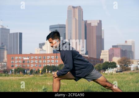 Young Hispanic sporty man in active wear doing lunge forward in green park with downtown on background Stock Photo