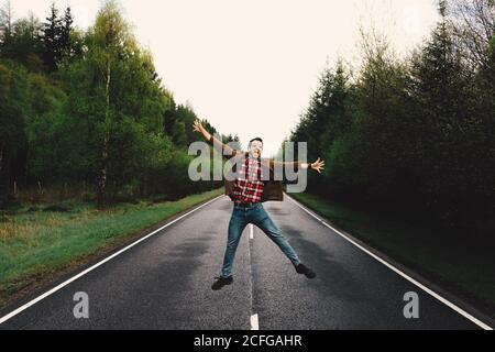 Young man jumping in solitude on remote road with lush green trees, Scotland Stock Photo