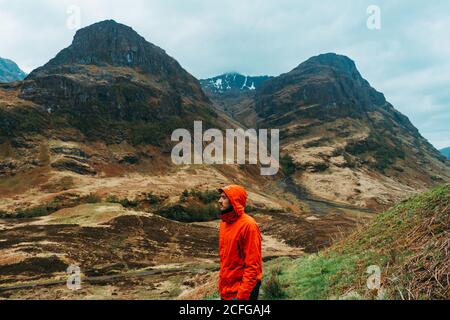 Side view of young man wearing a red raining coat hoodie while standing against picturesque mountains of Scotland Stock Photo