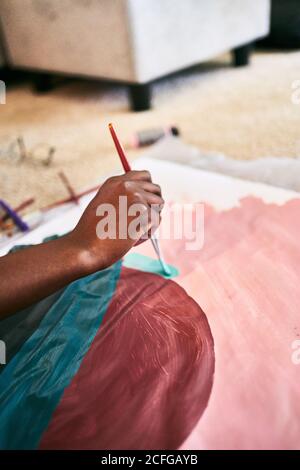 From above of crop anonymous black female with paintbrush drawing colorful picture on paper while sitting on floor at home Stock Photo
