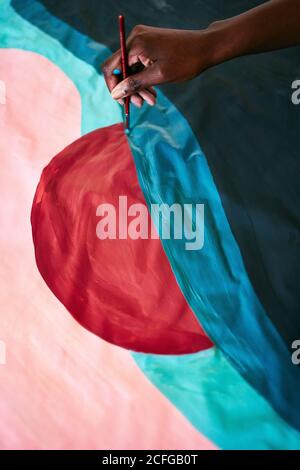 From above of crop anonymous black female with paintbrush drawing colorful picture on paper while sitting on floor at home Stock Photo