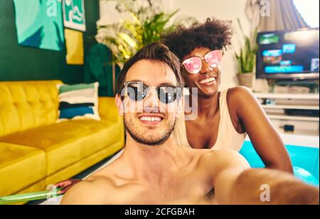 Cheerful multiracial couple wearing sunglasses sitting in inflatable pool and taking selfie while enjoying weekend Stock Photo