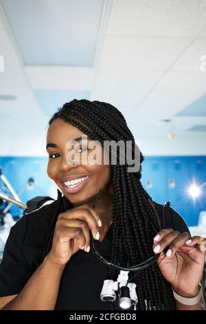Happy black Woman with braids smiling and looking at camera before putting on binocular goggles during work in contemporary laboratory Stock Photo