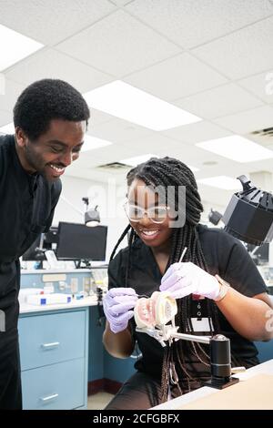Delighted African American female with braids smiling and using mouth mirror and probe to show coworker false teeth during work in lab Stock Photo