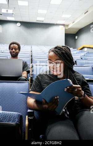 African American Woman in goggles reading notes while sitting near black man using laptop before lecture in modern auditorium Stock Photo