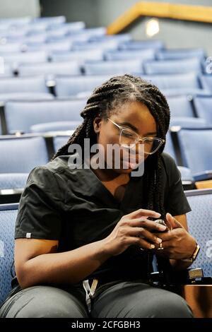 Black Woman sitting in auditorium and browsing smartphones during lesson in auditorium Stock Photo