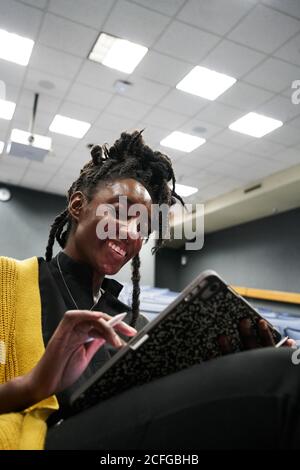 From below black Woman with dreadlocks smiling and using tablet while sitting in illuminated lecture hall during lesson Stock Photo