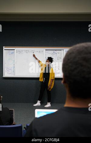 Full length African American Woman in casual clothes pointing at whiteboard and explaining data to audience during lecture in university Stock Photo