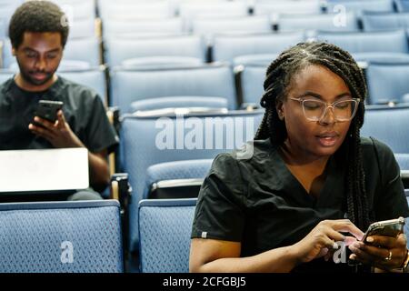 Black Woman with braids and African American man sitting in auditorium and browsing smartphones during lesson in auditorium Stock Photo