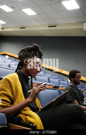 From below black Woman with dreadlocks smiling and using tablet while sitting in illuminated lecture hall during lesson Stock Photo