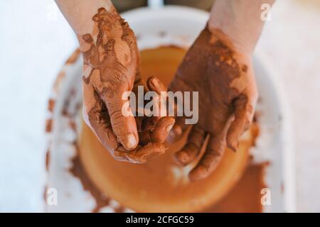From above of crop person hands in brown clay after making pottery against blurred special equipment in light modern workshop Stock Photo