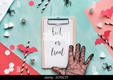 Trick or treat text on clipboard pad on layered paper background in mint green and red. Flat lay, Halloween party, hand in glove, confetti, drink stra Stock Photo