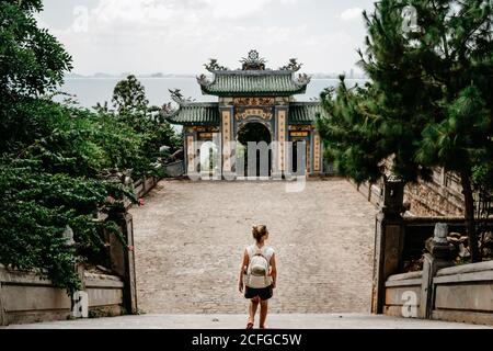 Back view of anonymous female tourist with backpack standing on stairs in front of arch of Buddhist temple in Vietnam Stock Photo