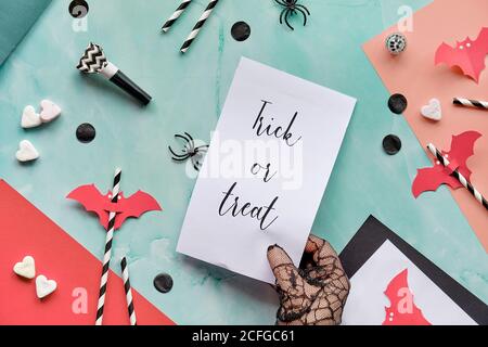 Trick or treat text on page held in gloved hand. Layered paper background in mint green and red. Top view, Halloween party decor, mirror balls, confet Stock Photo