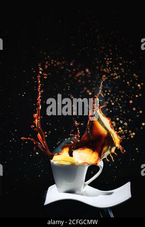 Falling slice of fresh lemon in splashing hot fresh coffee in white cup on white plate in dark room on black background Stock Photo