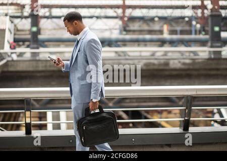 Handsome black businessman in coat and with phone walking down moving stairs while commuting. Stock Photo