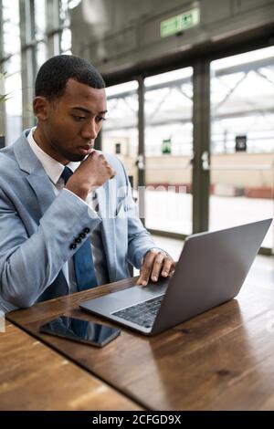 Young African-American man in blue suit sitting at wooden table and watching laptop in relax. Stock Photo