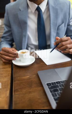 faceless businessman sitting at desk with cup of freshly brewed coffee and taking notes in pad with pen. Stock Photo