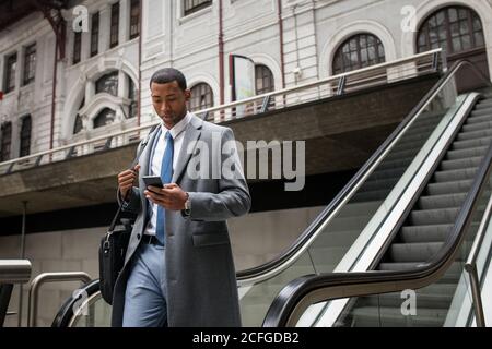 Handsome black businessman in coat and with phone walking down moving stairs while commuting. Stock Photo