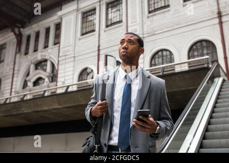 tHandsome black businessman in coat and with phone walking down moving stairs while commuting. Stock Photo