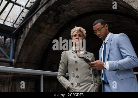 Laughing elegant man and Woman with papers and smartphone standing on bridge of railway station and chatting. Stock Photo