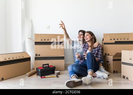 Young couple of man pointing up and Woman holding tablet and smiling sitting on floor near boxes and tools Stock Photo