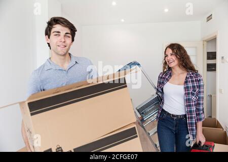 Young couple holding carton boxes and ladder in apartment and smiling Stock Photo